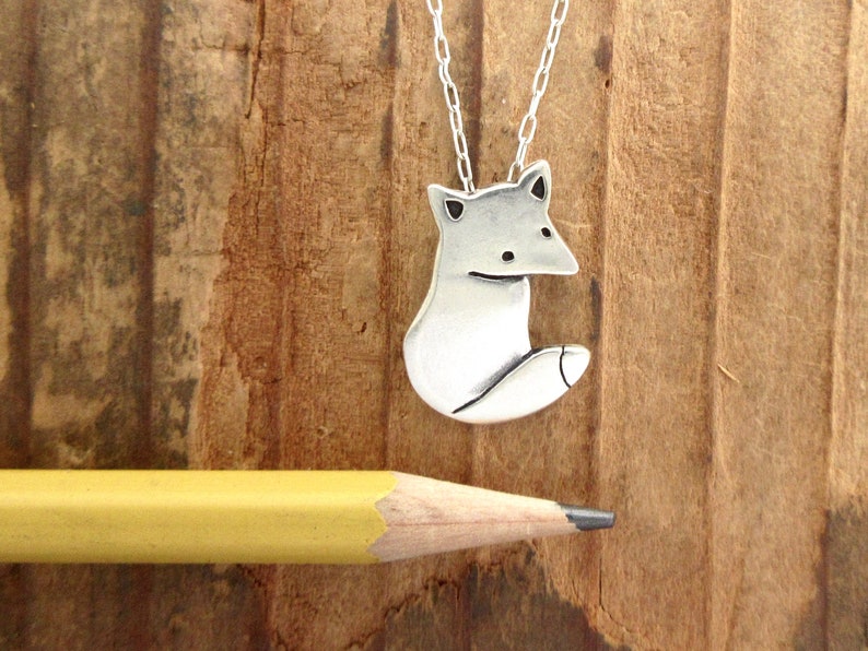 May include: A silver fox pendant necklace on a silver chain. The fox is sitting with its tail curled around its body. The necklace is on a wooden surface.