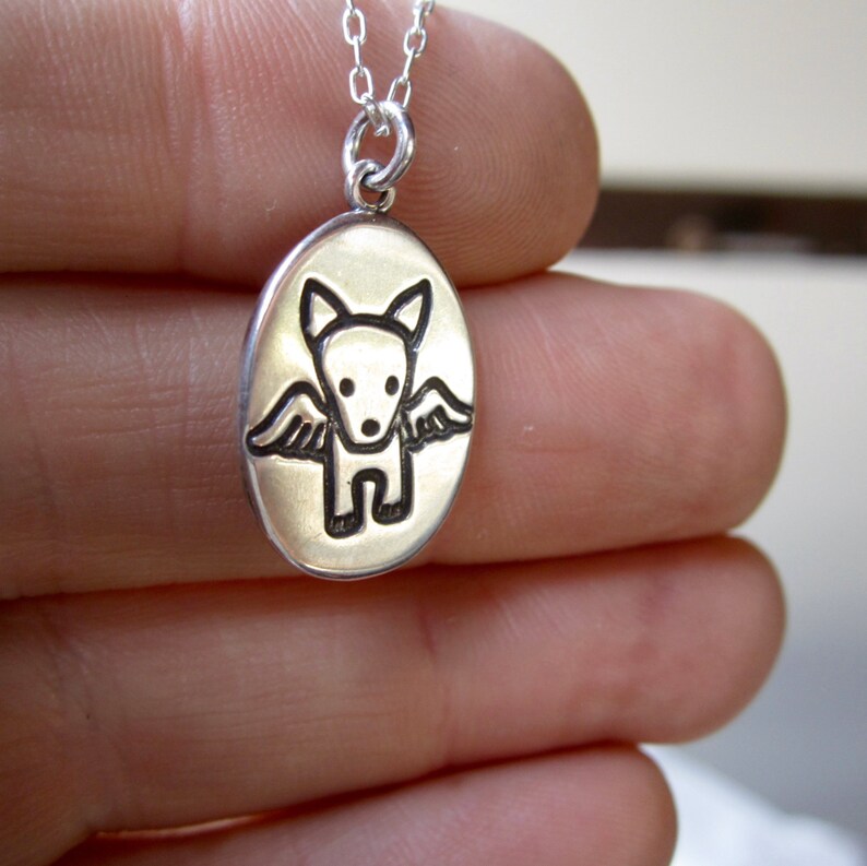 May include: Silver pendant necklace featuring an oval charm with a dog-like figure with wings. The dog is black, and the background is a light gold color. The necklace chain is delicate and silver.