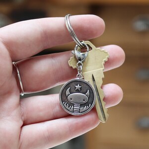 May include: Silver keyring with a round charm featuring a cartoon monster with a star on its head. The charm is attached to a silver key ring and a brass key.