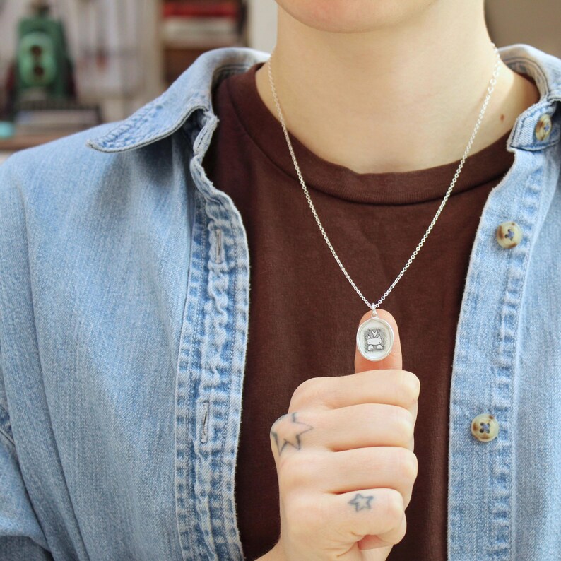 May include: A silver chain necklace with a small, round pendant featuring a detailed design. The pendant is being held by a person's thumb and forefinger.