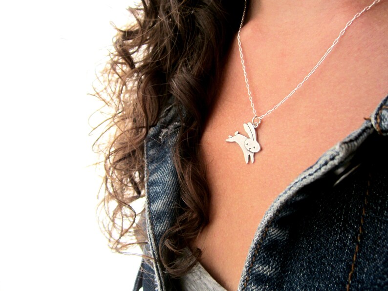 May include: A silver necklace featuring a delicate rabbit pendant. The rabbit is depicted in a forward-facing pose with its ears erect and tail tucked behind it.