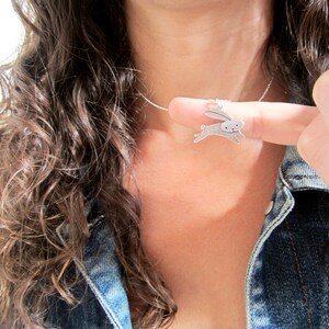 May include: A silver necklace with a small, silver rabbit pendant. The rabbit is facing right and has its ears up. The necklace is worn by a person with long, brown hair.