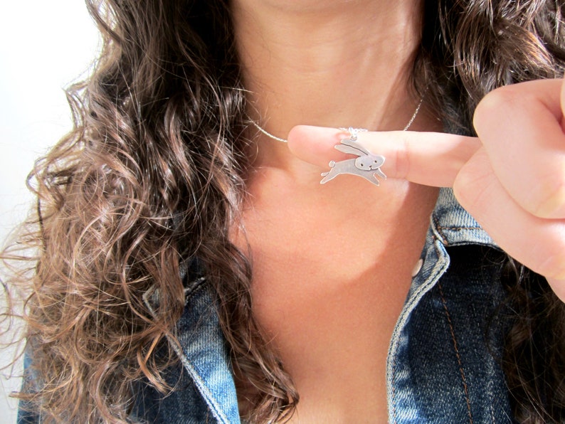 May include: A silver necklace with a small, silver rabbit pendant. The rabbit is facing right and has its ears up. The necklace is worn by a person with long, brown hair.