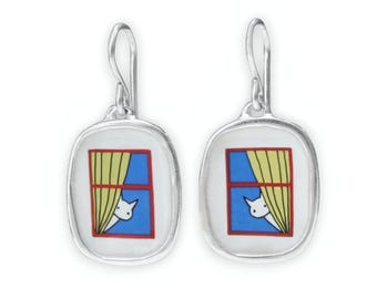 May include: A pair of silver dangle earrings with a white cat peeking out of a window with yellow curtains. The window is framed in red and blue.