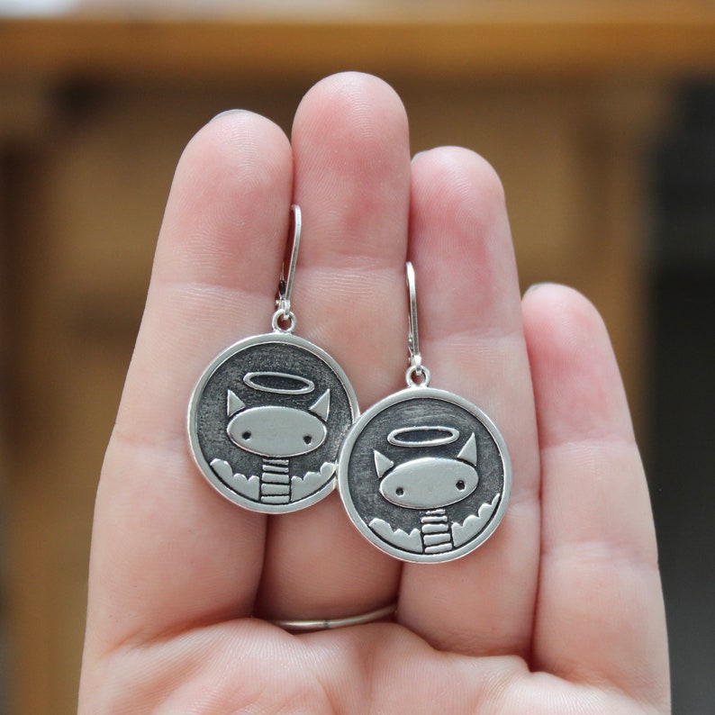 May include: A pair of silver earrings featuring a cat with a halo. The cat is depicted in a simple, cartoon-like style.