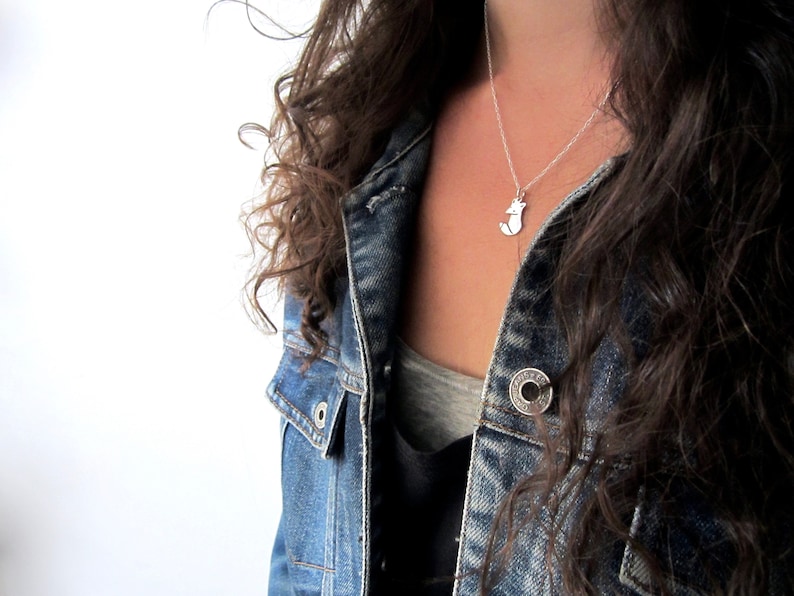 May include: A silver necklace with a small fox pendant. The necklace is worn by a person wearing a denim jacket.