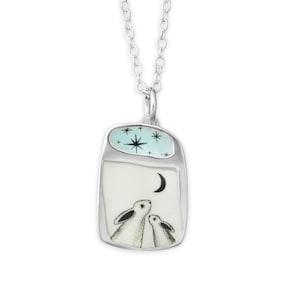 Peut inclure: Un collier pendentif argenté avec un pendentif rectangulaire. Le pendentif représente deux lapins blancs regardant un croissant de lune sous un ciel étoilé. La partie supérieure a un fond bleu clair avec des étoiles.