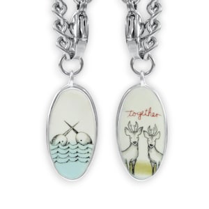 Könnte beinhalten: Zwei silberne Charm-Armbänder mit ovalen Porzellan-Anhängern. Ein Anhänger zeigt eine Schwarzweiß-Illustration von zwei Narwalen mit dem Text "together" in Rot. Der andere Anhänger zeigt eine Schwarzweiß-Illustration von zwei Hirschen.