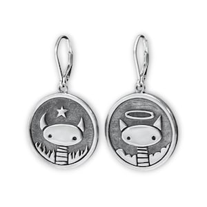 May include: A pair of silver dangle earrings with a round charm featuring a cartoon character with horns and a halo. One charm has a star above the character's head, and the other charm has a halo above the character's head.