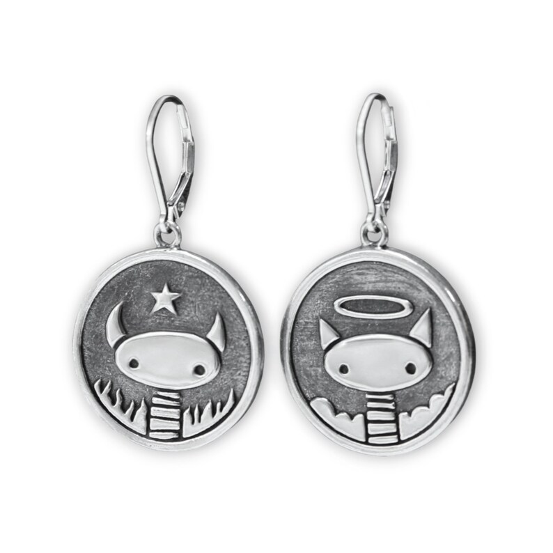 May include: A pair of silver dangle earrings with a round charm featuring a cartoon character with horns and a halo. One charm has a star above the character's head, and the other charm has a halo above the character's head.