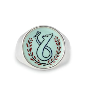 May include: A silver ring with a light blue enamel inlay featuring a stylized white rabbit and fox design. The design is surrounded by red and black leaves.