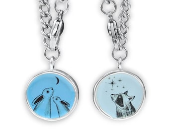 Puede incluir: Dos collares de plata con dijes de esmalte azul. Un dije presenta dos conejos mirando hacia arriba a una luna creciente. El otro dije presenta un mapache mirando hacia arriba a una estrella.