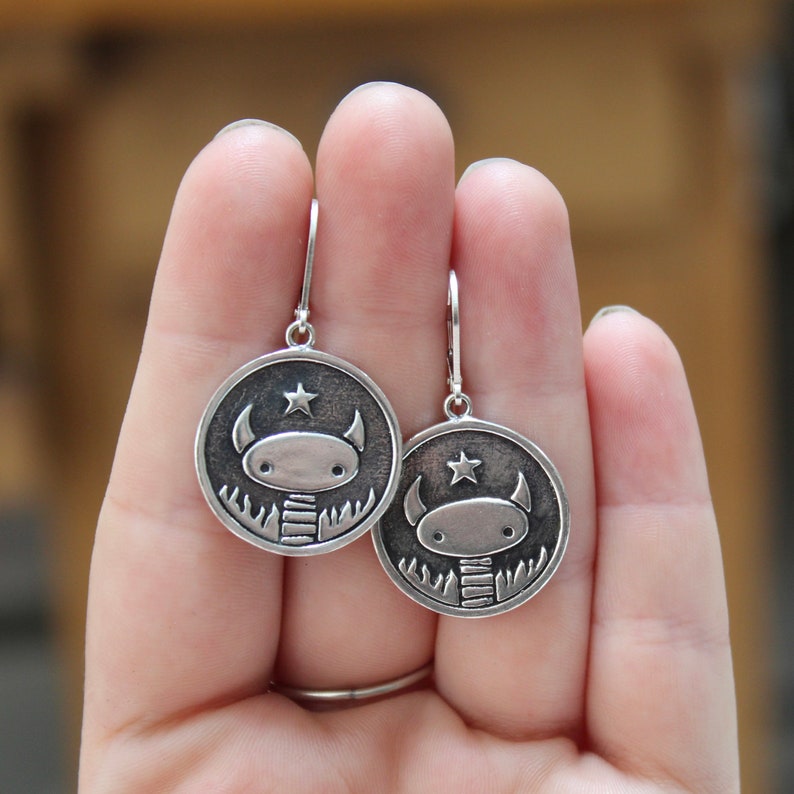 May include: A pair of silver dangle earrings featuring a cartoon monster with a star on its head. The monster is wearing a striped scarf and is surrounded by a circle of leaves.