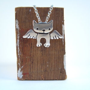 Puede incluir: Collar de plata con un colgante de un gato con alas. El gato est&aacute; de frente con las alas extendidas. El collar est&aacute; en un soporte de madera.