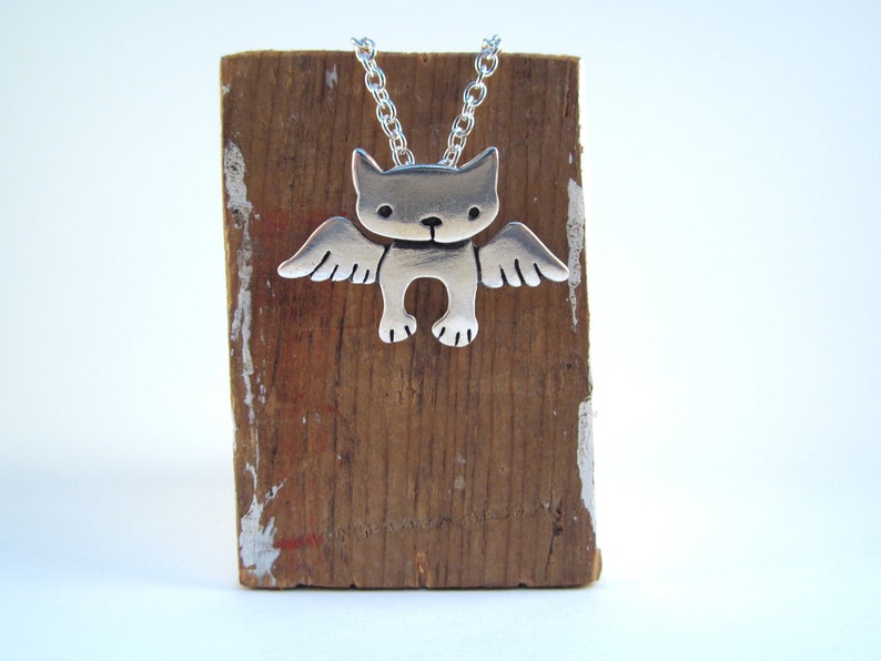 Puede incluir: Collar de plata con un colgante de un gato con alas. El gato est&aacute; de frente con las alas extendidas. El collar est&aacute; en un soporte de madera.