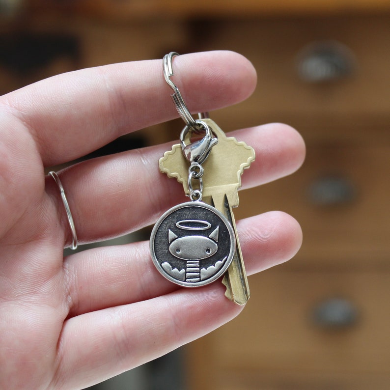 May include: A silver keyring with a small, round charm featuring a cartoon cat with a halo. The charm is attached to a silver key ring and a brass key.