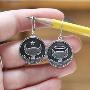 May include: A pair of silver earrings with black and white illustrations of a devil and an angel. The devil has horns and a star above its head, while the angel has a halo and wings.