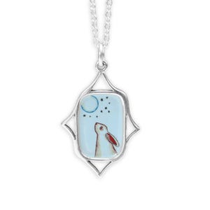 May include: A silver necklace with a rectangular pendant featuring a white rabbit looking up at a blue crescent moon and stars on a light blue background.