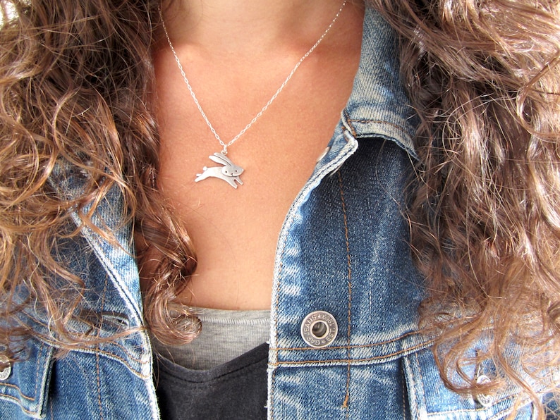 May include: A silver necklace featuring a small, stylised rabbit pendant. The necklace is worn by a person with long, brown hair and a denim jacket.