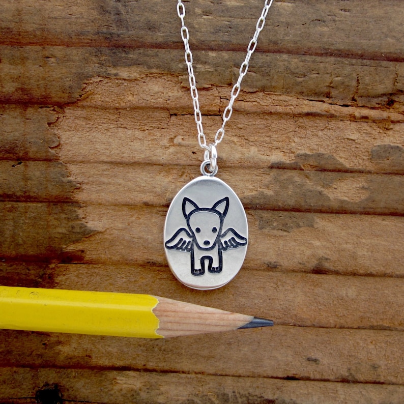 May include: A silver pendant necklace featuring an oval charm with a stylized dog design with wings. The dog is black and the wings are detailed. The necklace chain is delicate and silver. A yellow pencil is in the foreground.