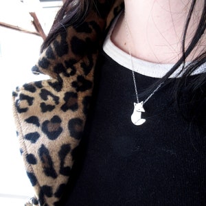 May include: A silver necklace with a small fox pendant. The necklace is worn by a person with long black hair and a leopard print jacket.