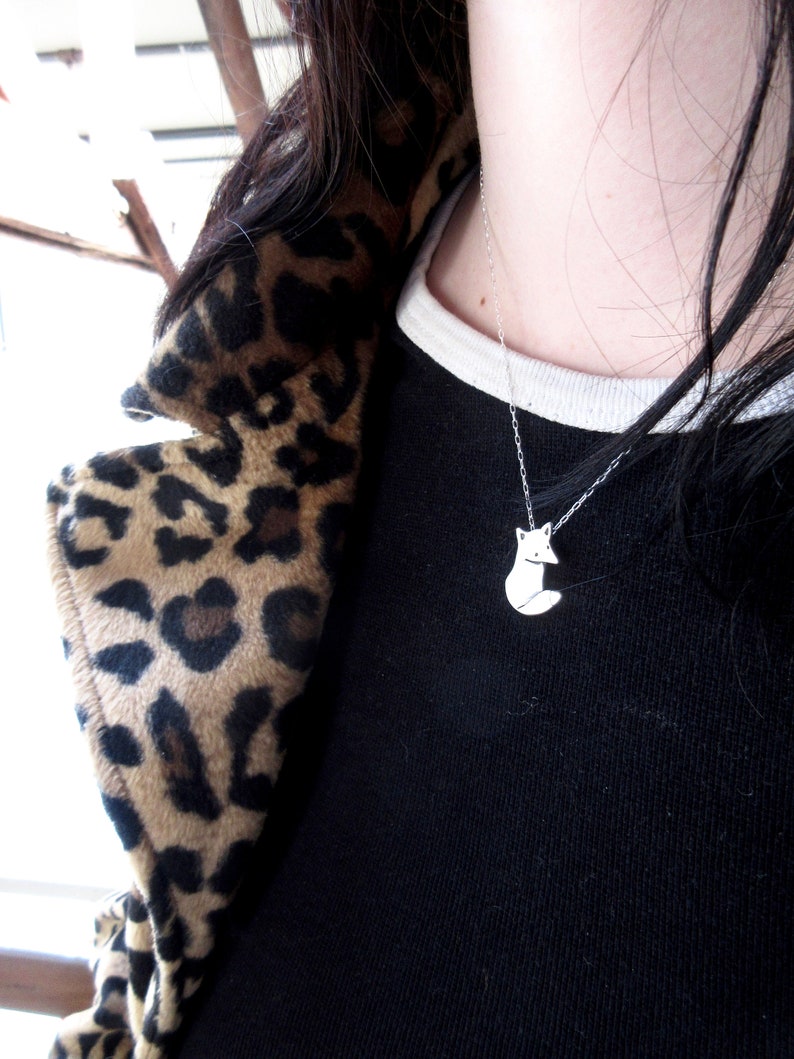 May include: A silver necklace with a small fox pendant. The necklace is worn by a person with long black hair and a leopard print jacket.