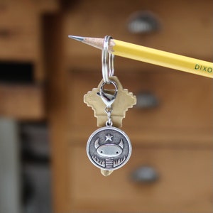 May include: A silver keychain with a small, round charm featuring a cartoon creature with a star on its head. The charm is attached to a silver key ring and a silver key.