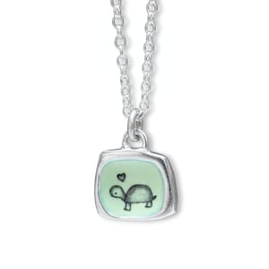 May include: A silver necklace with a square pendant. The pendant has a light green background with a black and white turtle illustration and a small heart. The necklace is displayed on a white background.