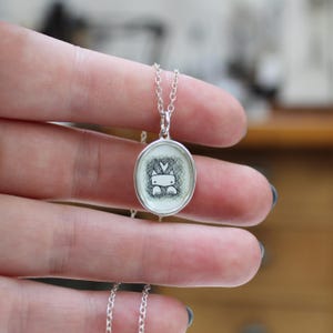 May include: A silver chain necklace with a round pendant featuring a black and white illustration of a cat with a heart on its head.