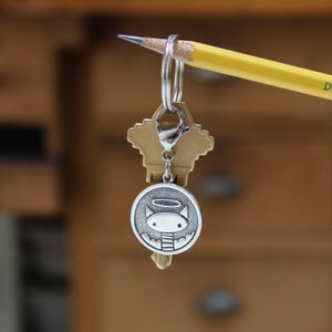 May include: A silver key chain with a round charm featuring a cartoon cat with a halo. The charm is attached to a silver key ring and a brass key.