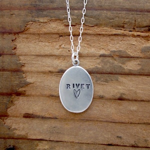 May include: Silver pendant necklace with an oval pendant. The pendant has the word "RIVET" stamped on it, with a heart symbol below. The necklace chain is also silver and hangs against a wooden background.