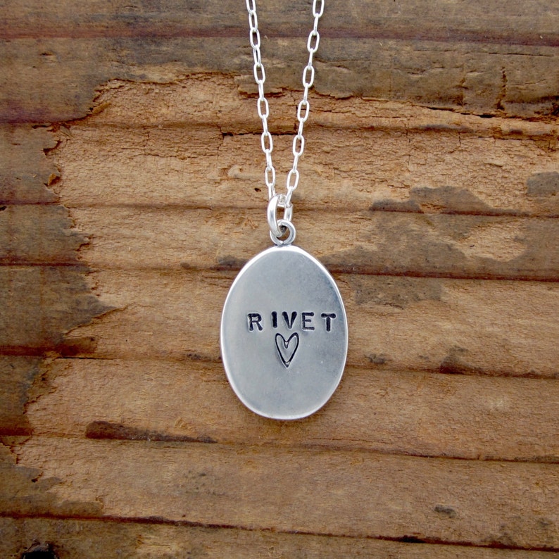 May include: Silver pendant necklace with an oval pendant. The pendant has the word "RIVET" stamped on it, with a heart symbol below. The necklace chain is also silver and hangs against a wooden background.