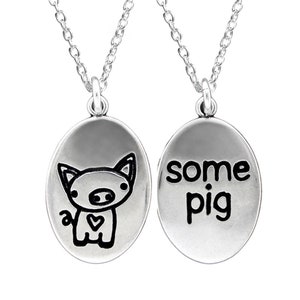 May include: A silver necklace with two oval pendants. One pendant features a cartoon pig with a heart on its chest. The other pendant says "some pig".
