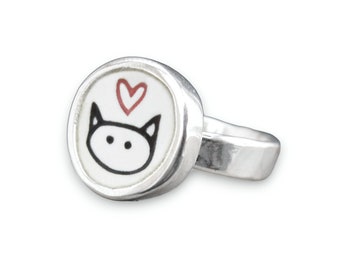 May include: A silver ring with a white enamel cat face and a red heart on top. The ring is a simple band.