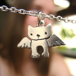 Puede incluir: Un collar de plata con un dije de un gato con alas. El gato est&aacute; de frente, con los ojos cerrados y tiene una peque&ntilde;a nariz y boca.