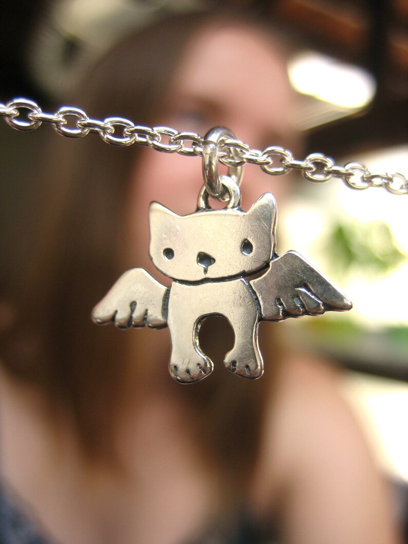 Puede incluir: Un collar de plata con un dije de un gato con alas. El gato est&aacute; de frente, con los ojos cerrados y tiene una peque&ntilde;a nariz y boca.