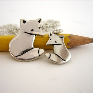 May include: Two silver fox charms, one larger than the other, on a silver chain. The larger charm is hanging from a yellow pencil.
