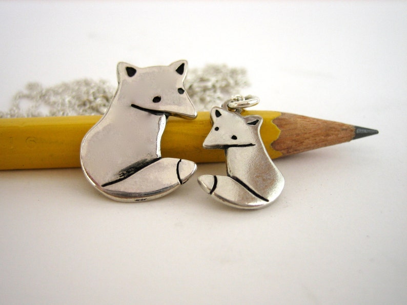 May include: Two silver fox charms, one larger than the other, on a silver chain. The larger charm is hanging from a yellow pencil.