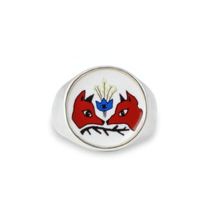 May include: A silver ring with a white enamel top featuring a stylized illustration of two red fox heads facing each other with a blue flower in between.