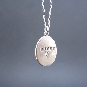 May include: A silver pendant necklace with an oval-shaped charm. The charm has the word "RIVET" stamped on it, with a small heart symbol below. The necklace chain is also silver.