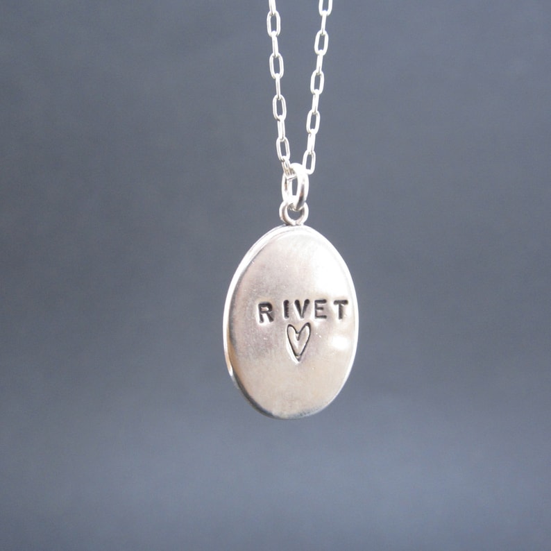May include: A silver pendant necklace with an oval-shaped charm. The charm has the word "RIVET" stamped on it, with a small heart symbol below. The necklace chain is also silver.