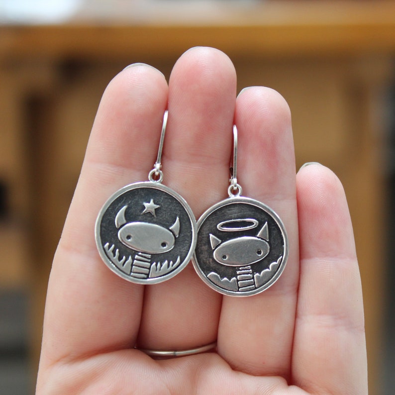 May include: A pair of silver dangle earrings with a black and white design. One earring features a cartoon angel with a halo and the other earring features a cartoon devil with a star.