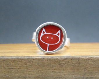 May include: A silver ring with a red enamel inlay featuring a simple white line drawing of a cat face.