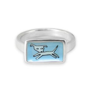 May include: A silver ring featuring a rectangular face with a light blue enamel inlay. The center of the inlay displays a simple black line drawing of a dog in a running or jumping pose. The band of the ring is smooth, polished, and metallic.