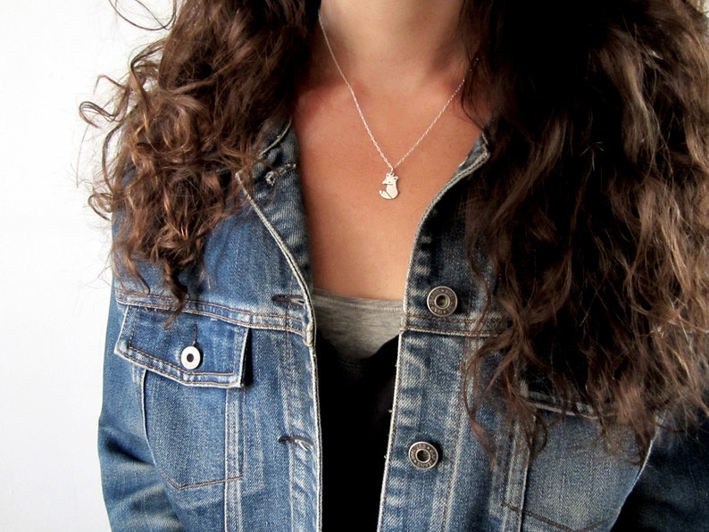 May include: A silver necklace with a small fox pendant. The necklace is worn by a person with long brown curly hair and a denim jacket.