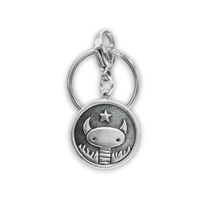 May include: Silver keyring with a round charm featuring a cartoon monster with horns and a star above its head. The monster is surrounded by flames.