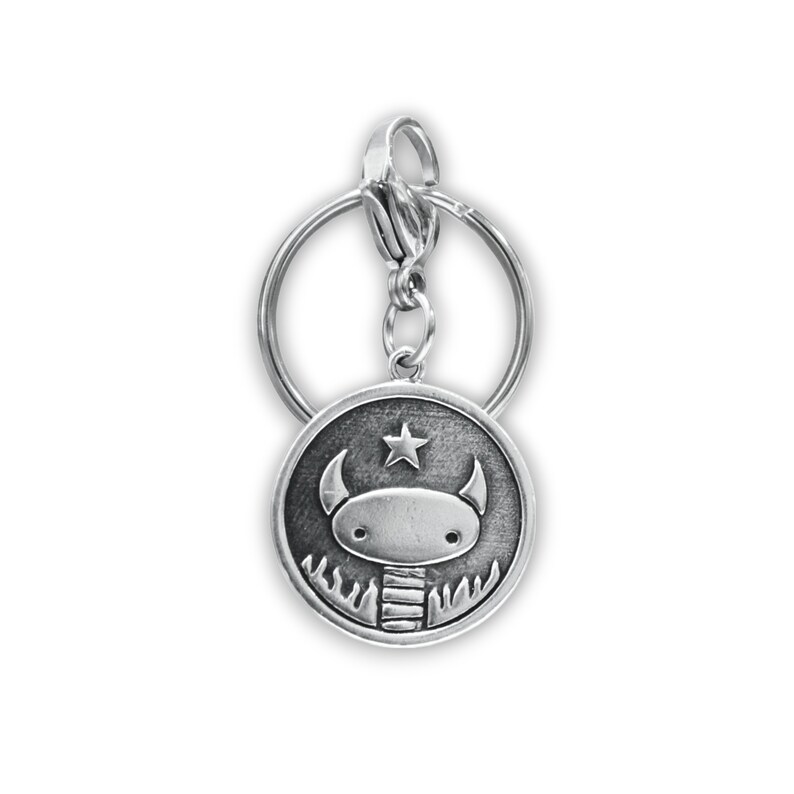 May include: Silver keyring with a round charm featuring a cartoon monster with horns and a star above its head. The monster is surrounded by flames.