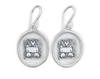 May include: A pair of silver dangle earrings with a white cat illustration in a round silver frame.