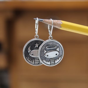 May include: Silver earrings with a circular design. One earring features a cartoon devil with horns and a star, the other earring features a cartoon angel with a halo and wings.