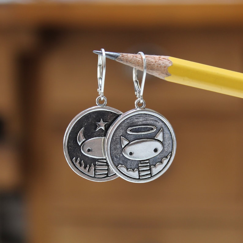 May include: Silver earrings with a circular design. One earring features a cartoon devil with horns and a star, the other earring features a cartoon angel with a halo and wings.